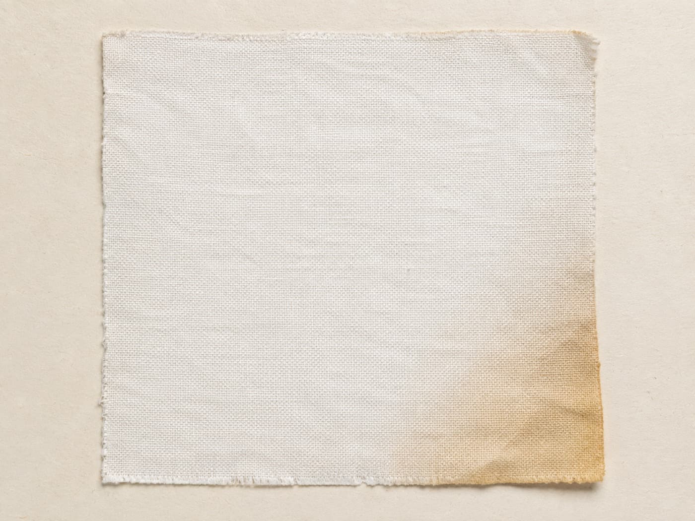 A square swatch of white cotton fabric on warm off-white paper, one corner slightly aged.