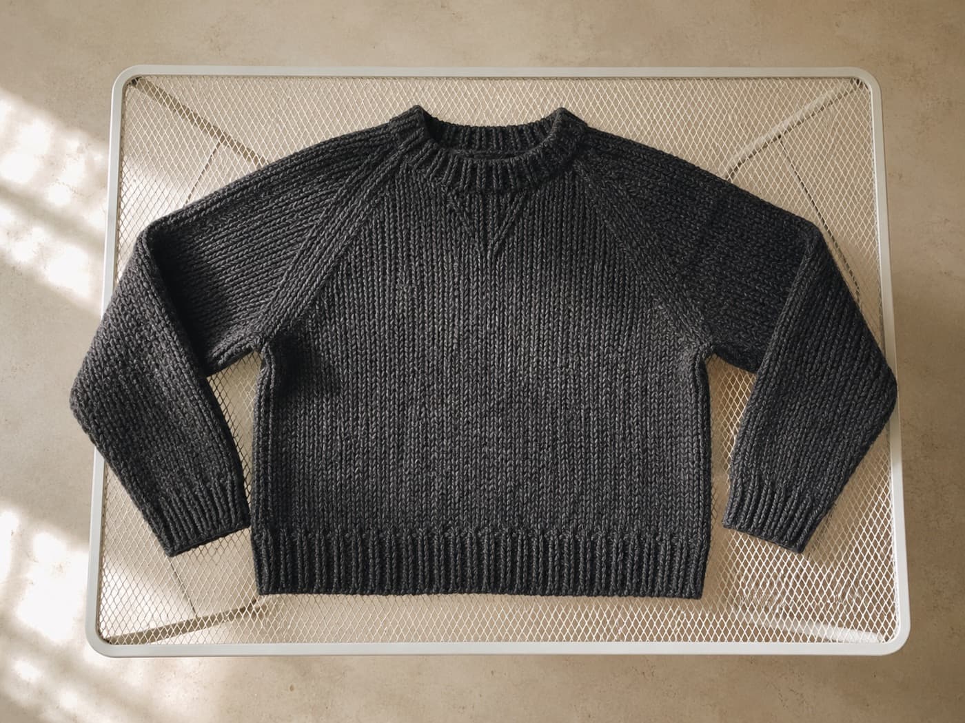 A charcoal wool knit sweater laid flat on a white mesh drying rack in daylight.
