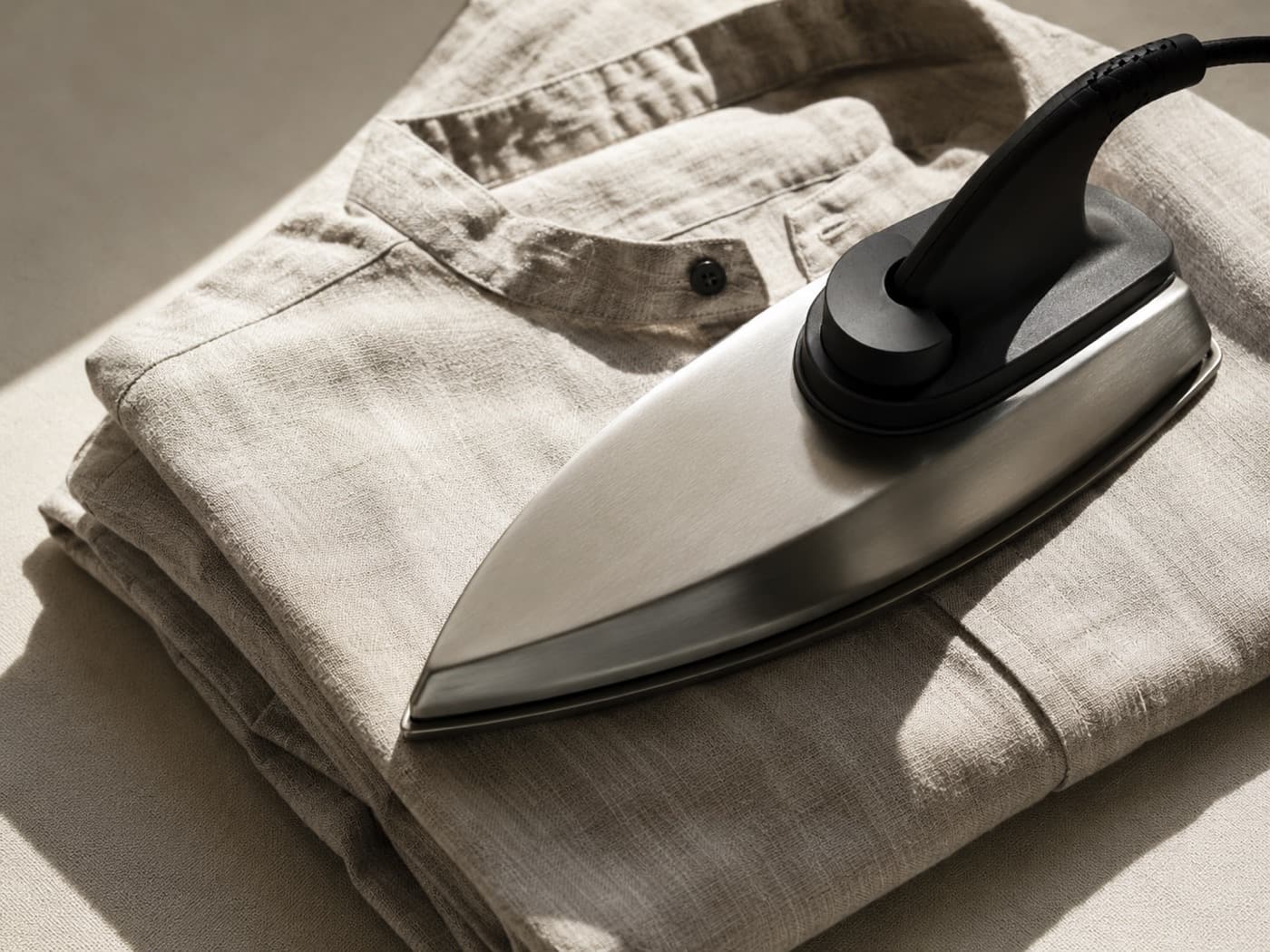 A matte steel iron soleplate resting on a folded natural linen shirt in raking side light.