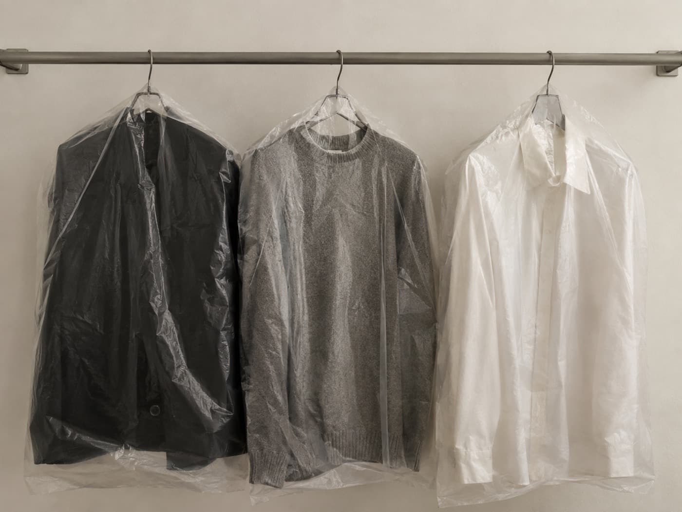 Three garments sealed in translucent protective film hanging on a plain steel rail.