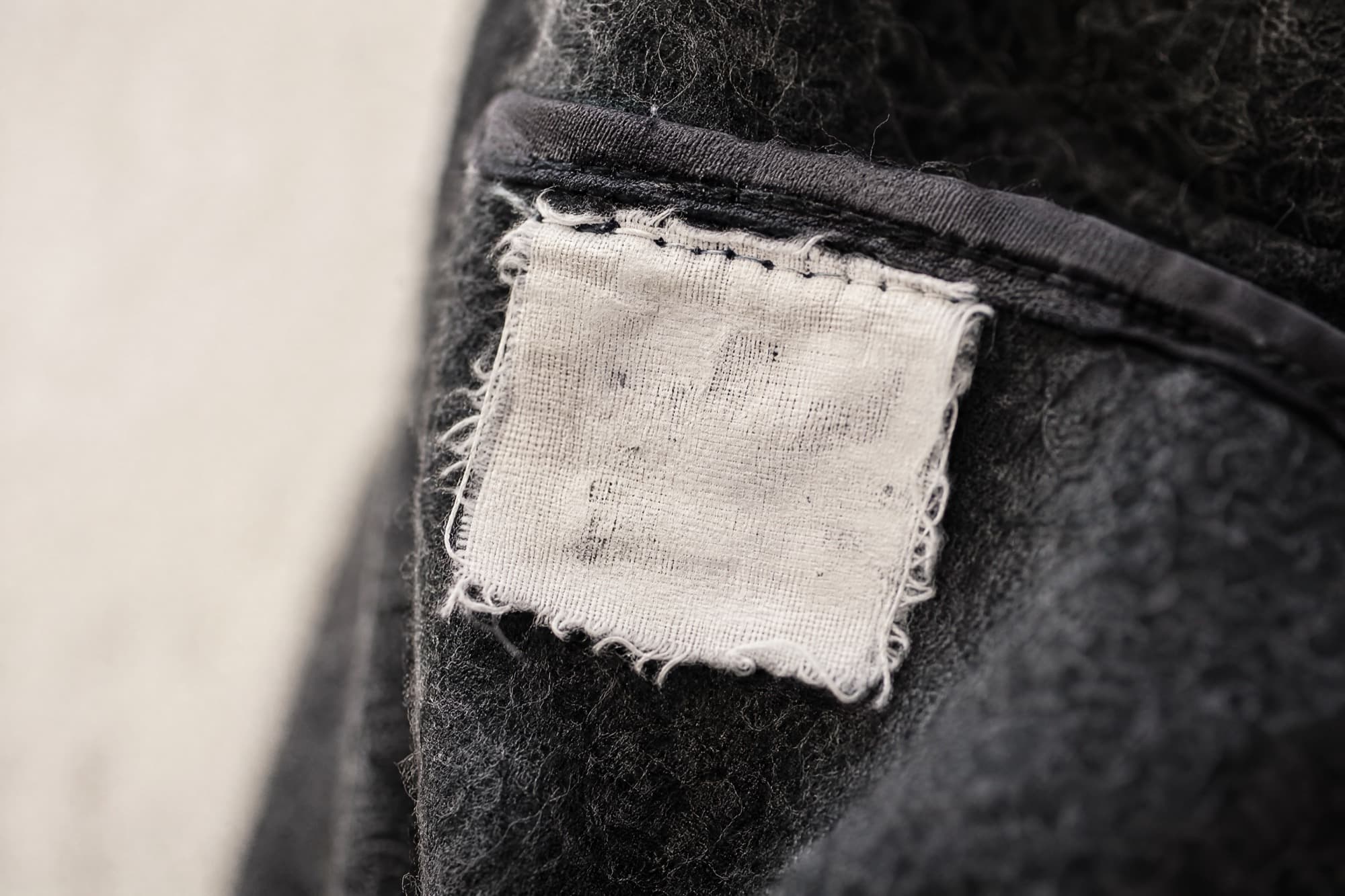 Extreme close-up of a small white care label stitched into a charcoal wool seam. Its printed markings are worn away and unreadable.